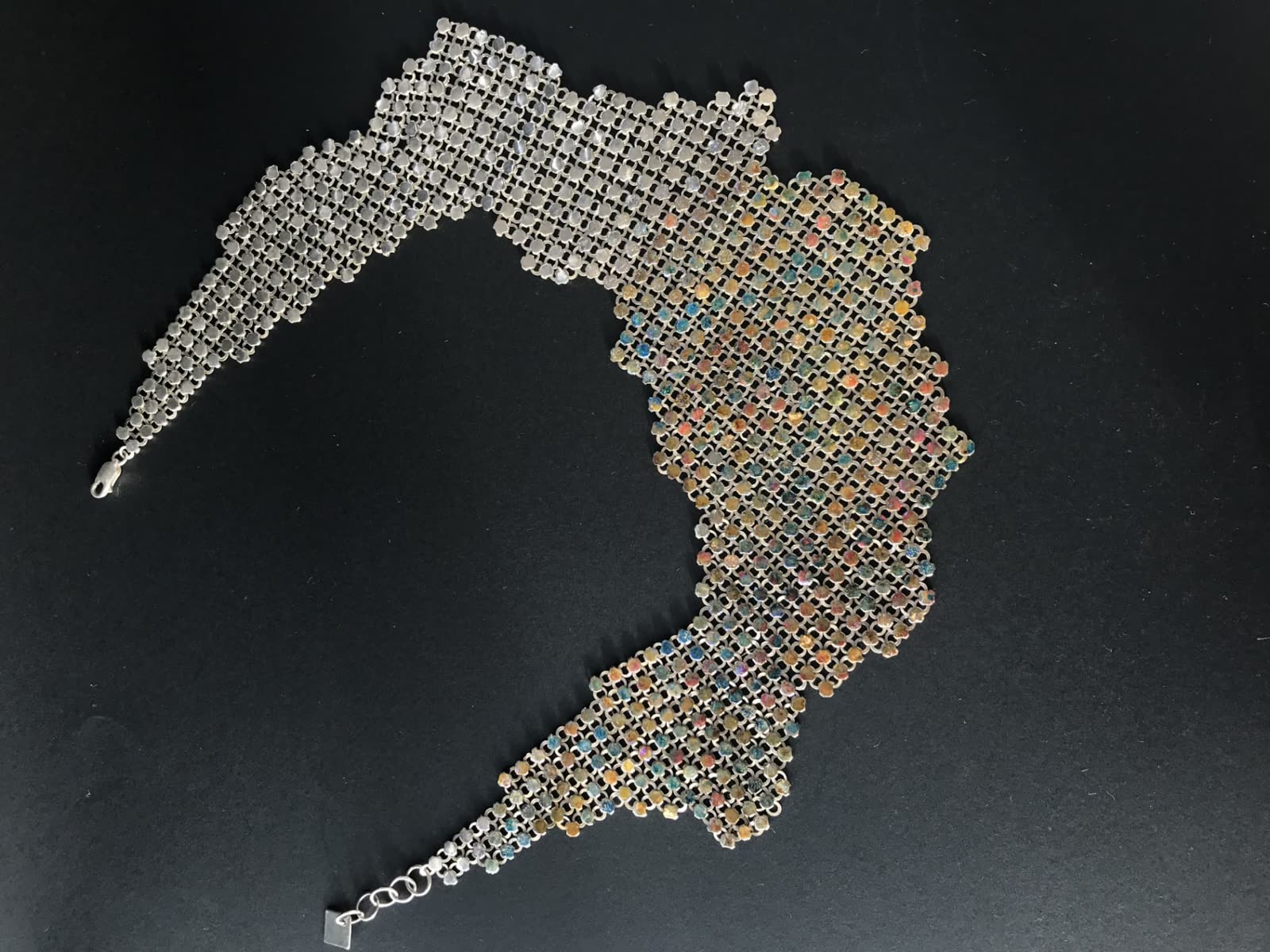 Collar de malla metálica tejida a mano con secciones esmaltadas en tonos ocre, azul y rosa, extendido sobre fondo negro