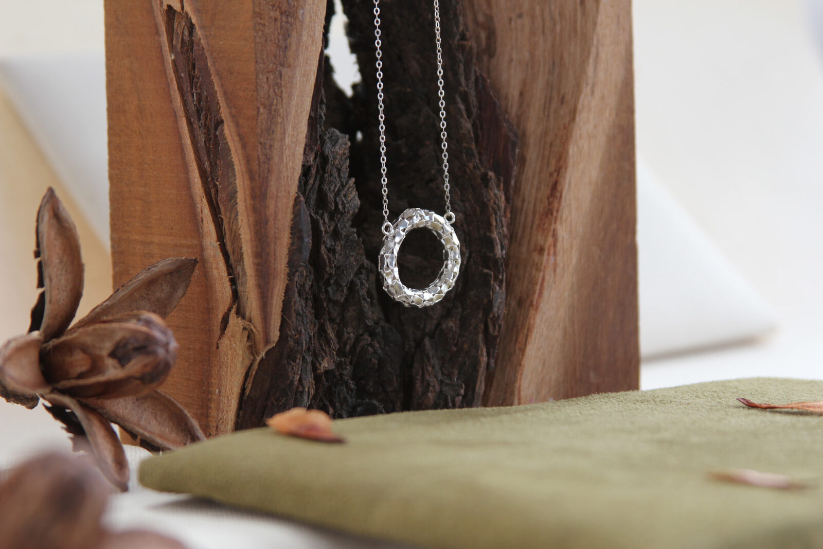 Collar con dije circular texturado en plata suspendido frente a un tronco de madera, junto a gamuza verde y cápsulas secas