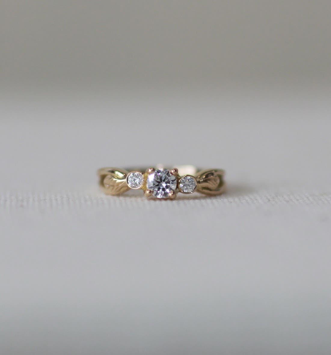 Anillo Tres Gracias en oro con diamante central y dos diamantes laterales, sobre fondo claro y mucho vacío alrededor