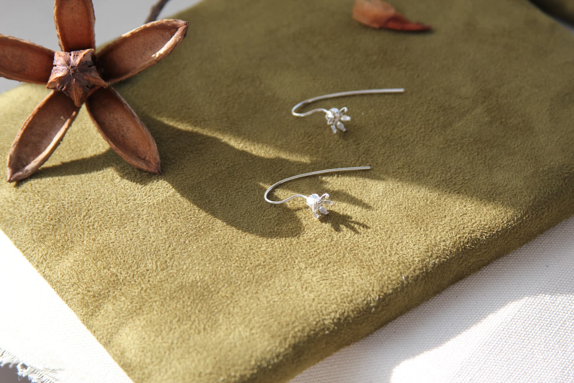 Par de aretes de gancho con flor de plata sobre gamuza verde oliva junto a una cápsula seca