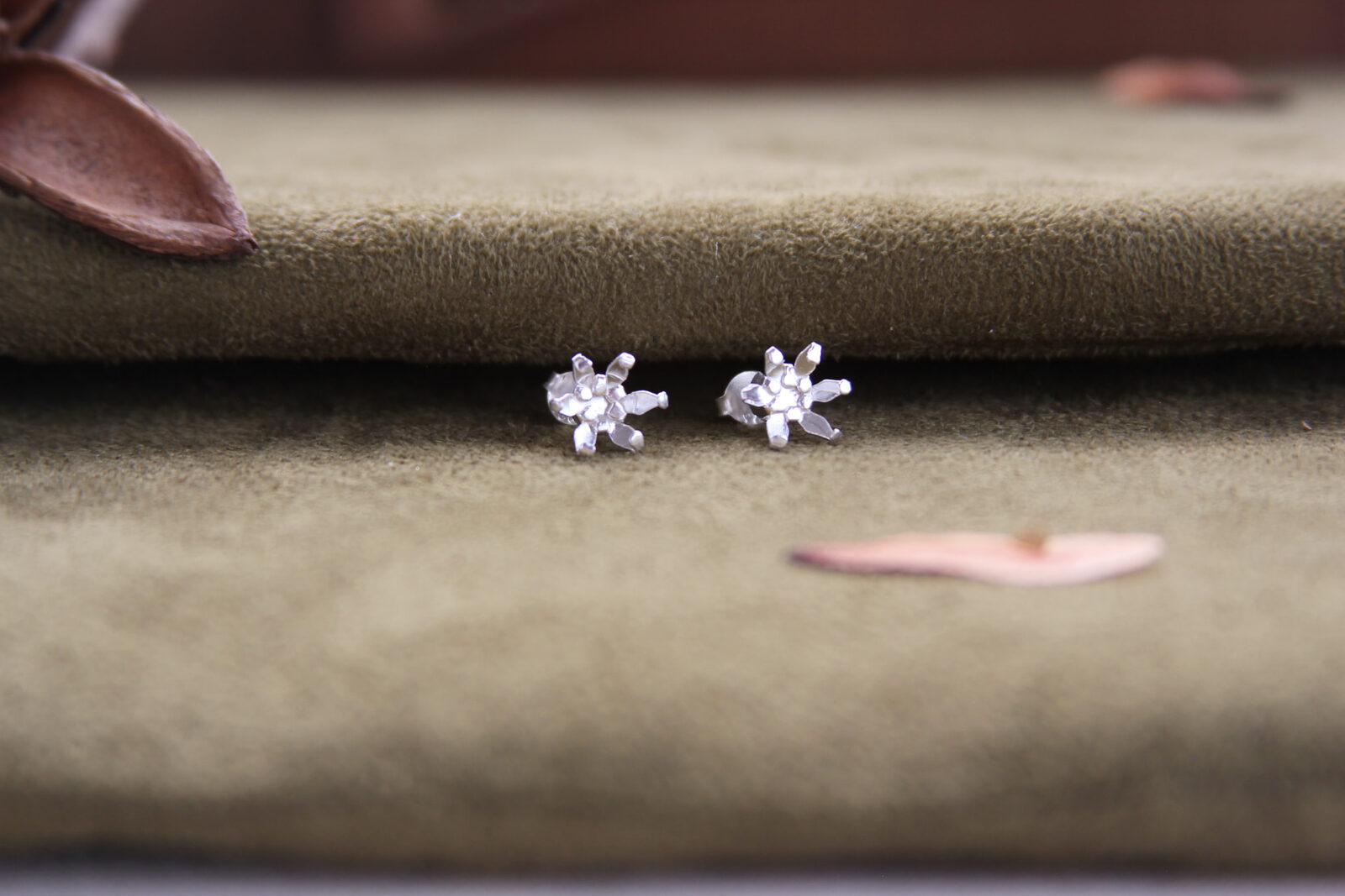 Topos en forma de estrella de plata sobre gamuza verde oliva, con pétalos secos y luz suave