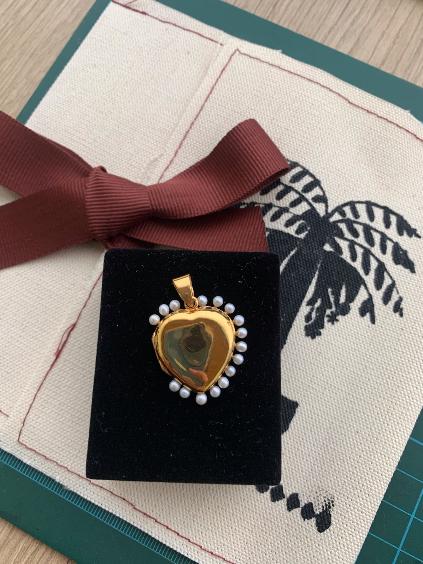 Relicario en forma de corazón con borde de perlas, en baño de oro, presentado en su estuche de terciopelo negro sobre lino con cinta vinotinto