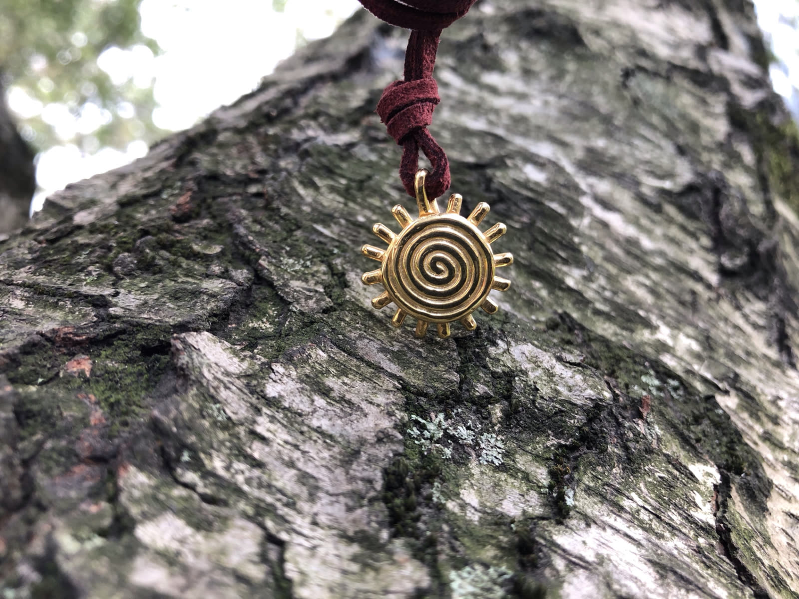 Amuleto solar en metal dorado con espiral central, colgado de un cordón de gamuza vinotinto sobre la corteza de un árbol