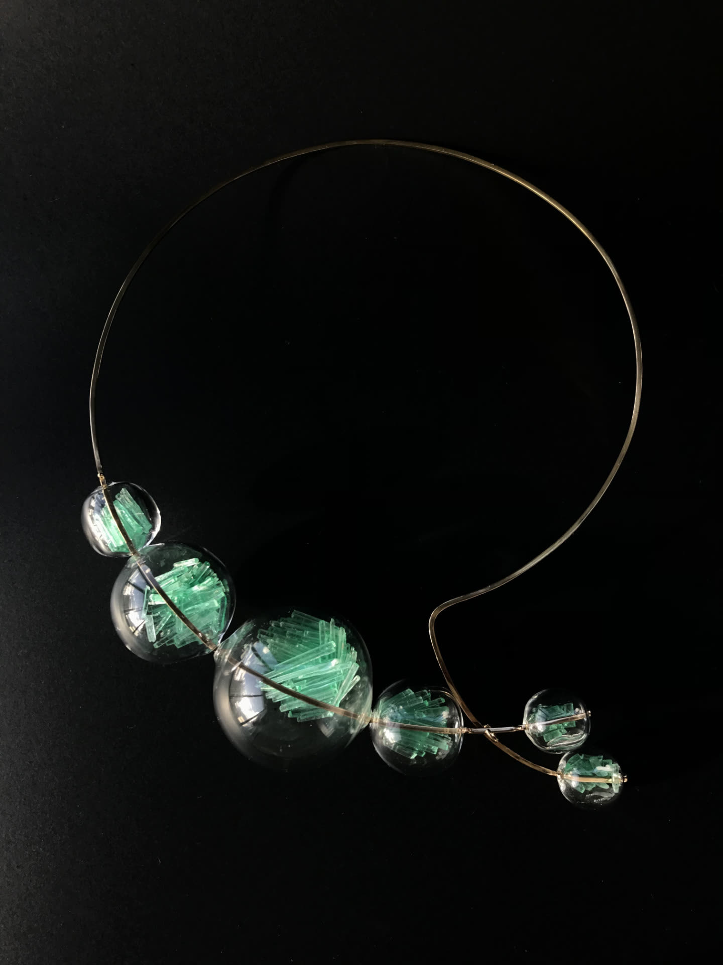 Collar rígido en metal con esferas de vidrio transparente que contienen cristales verdes, sobre fondo negro