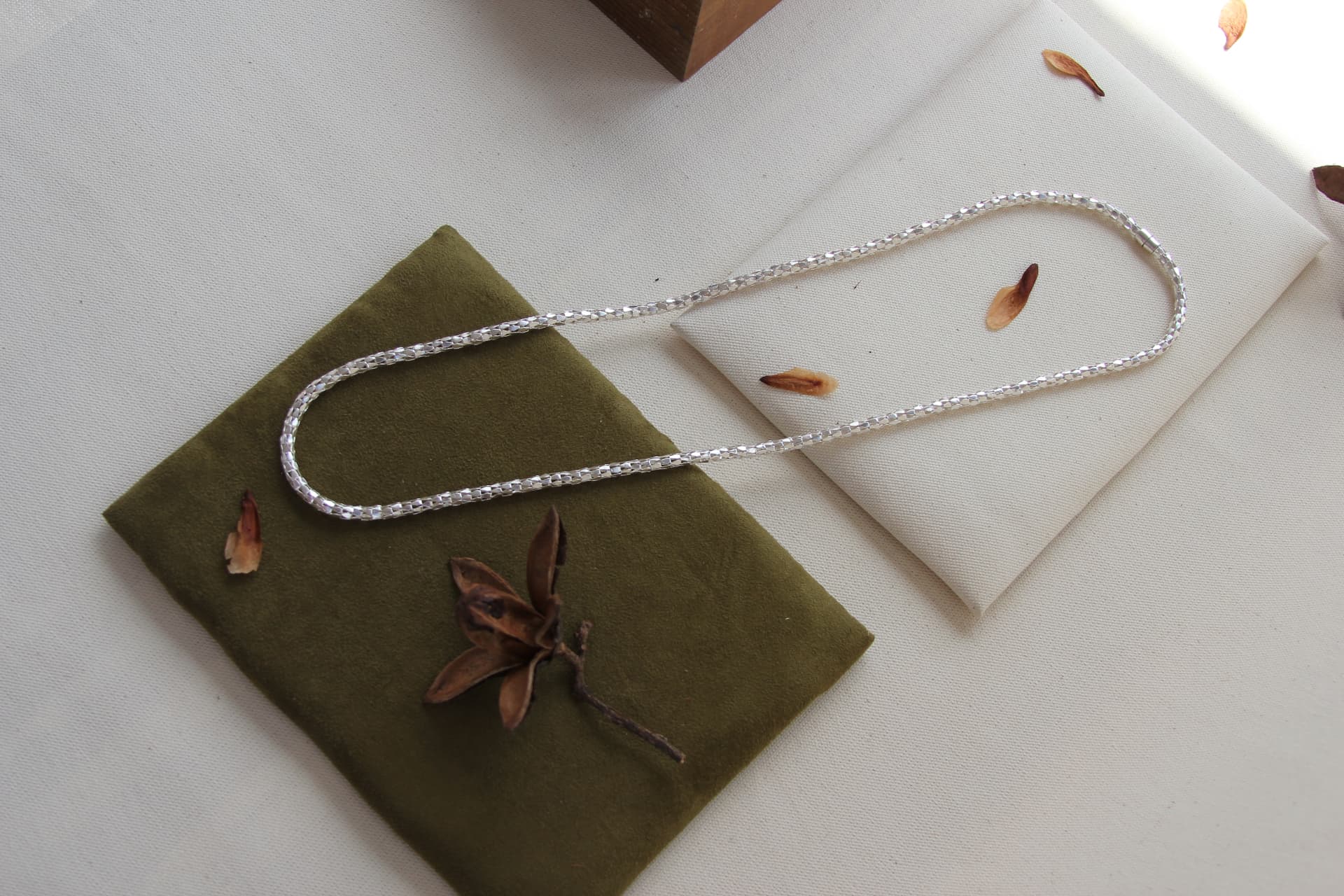 Collar de cadena facetada en plata sobre gamuza verde oliva y lino natural, con pétalos secos