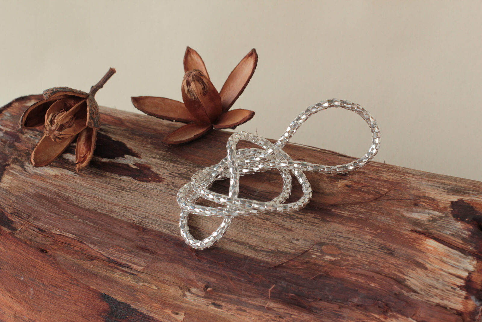 Brazalete anudado de cadena facetada en plata sobre un tronco de madera con cápsulas secas
